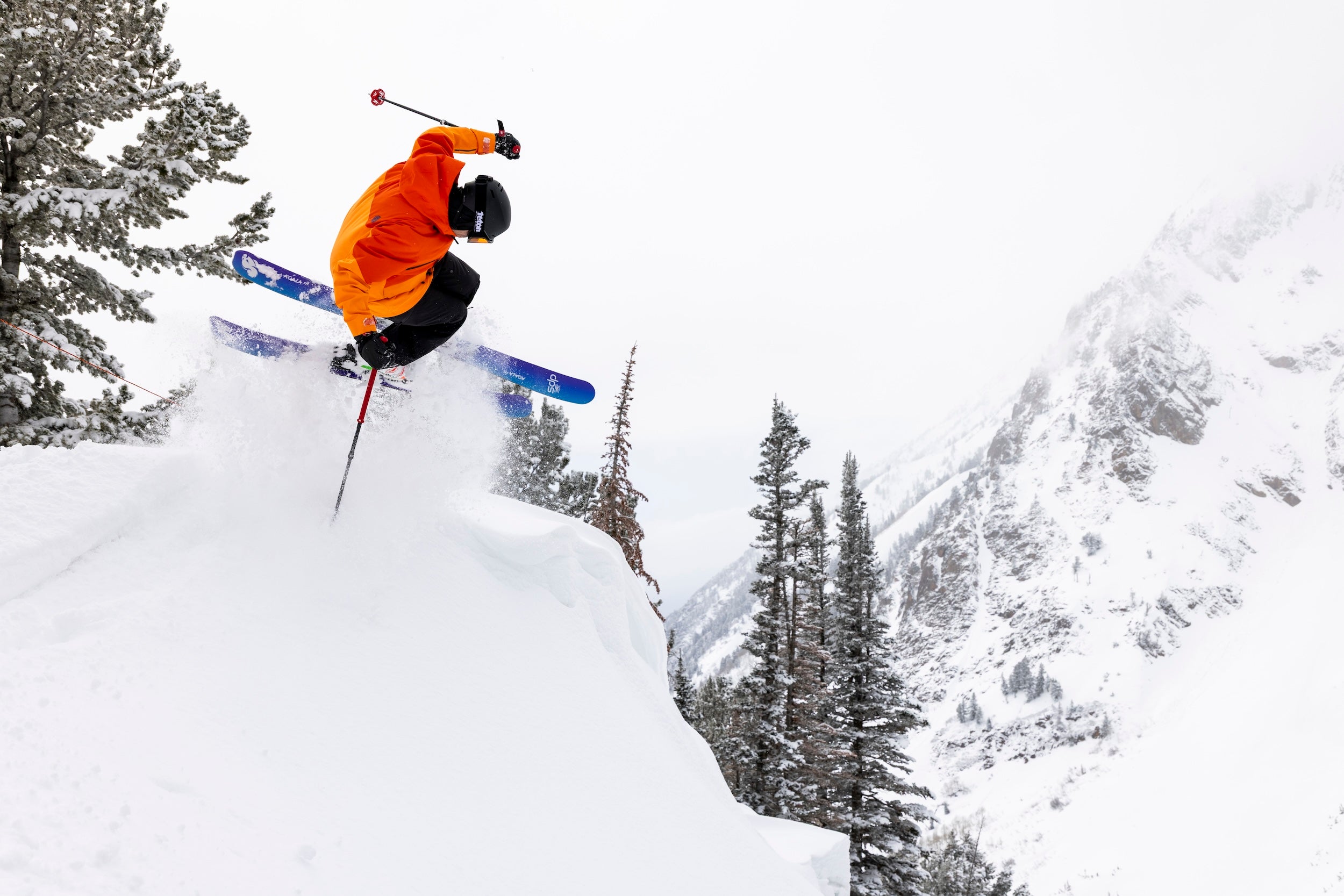 Skier in orange jacket performing a jump on a snowy mountain with trees in the background